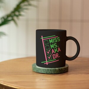 Puede incluir: Una taza de cer&aacute;mica negra con las palabras "MISS MS. AKA DR." en verde y rosa, con marcas de verificaci&oacute;n junto a "AKA" y "DR.". La taza est&aacute; sobre un posavasos de m&aacute;rmol verde en una mesa de madera.