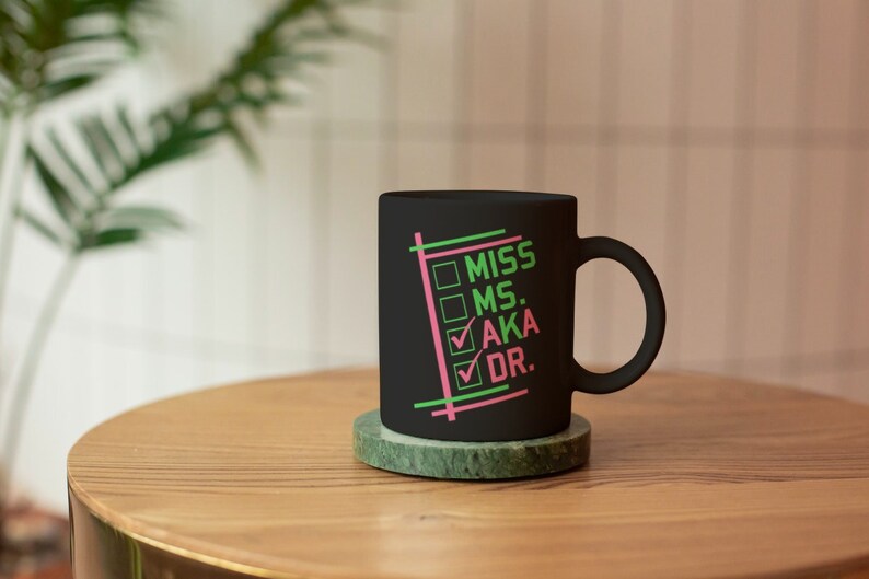 Puede incluir: Una taza de cer&aacute;mica negra con las palabras "MISS MS. AKA DR." en verde y rosa, con marcas de verificaci&oacute;n junto a "AKA" y "DR.". La taza est&aacute; sobre un posavasos de m&aacute;rmol verde en una mesa de madera.