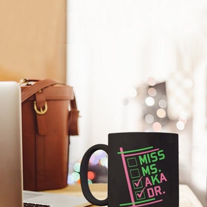 Puede incluir: Taza de cer&aacute;mica negra con las palabras "MISS MS. AKA DR." en verde y rosa. La taza tiene un asa negra y un dise&ntilde;o de casillas marcadas. Un port&aacute;til y una bolsa de cuero marr&oacute;n est&aacute;n en el fondo.