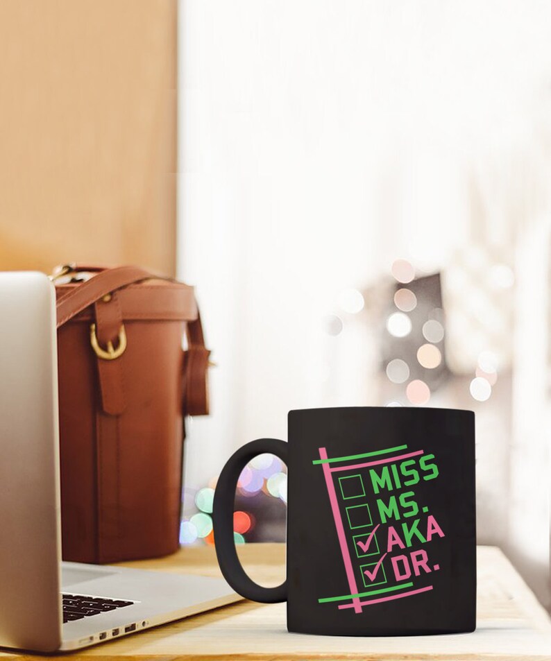 Puede incluir: Taza de cer&aacute;mica negra con las palabras "MISS MS. AKA DR." en verde y rosa. La taza tiene un asa negra y un dise&ntilde;o de casillas marcadas. Un port&aacute;til y una bolsa de cuero marr&oacute;n est&aacute;n en el fondo.