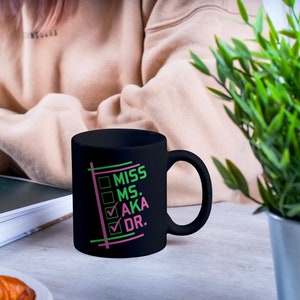 Puede incluir: Una taza de cer&aacute;mica negra con un asa curvada. La taza presenta las palabras "MISS MS. AKA DR." en verde y rosa, con marcas de verificaci&oacute;n junto a "AKA" y "DR." La taza est&aacute; sobre una superficie gris claro.
