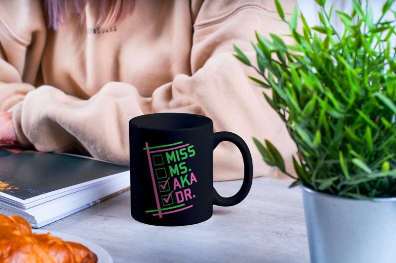 Puede incluir: Una taza de cer&aacute;mica negra con un asa curvada. La taza presenta las palabras "MISS MS. AKA DR." en verde y rosa, con marcas de verificaci&oacute;n junto a "AKA" y "DR." La taza est&aacute; sobre una superficie gris claro.