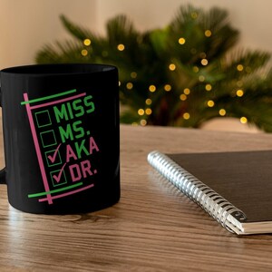 Puede incluir: Taza de cer&aacute;mica negra con asa blanca. La taza presenta las palabras "MISS, MS., AKA, DR." en verde y rosa, con marcas de verificaci&oacute;n junto a "AKA" y "DR.". Un cuaderno y un &aacute;rbol de Navidad est&aacute;n en el fondo.