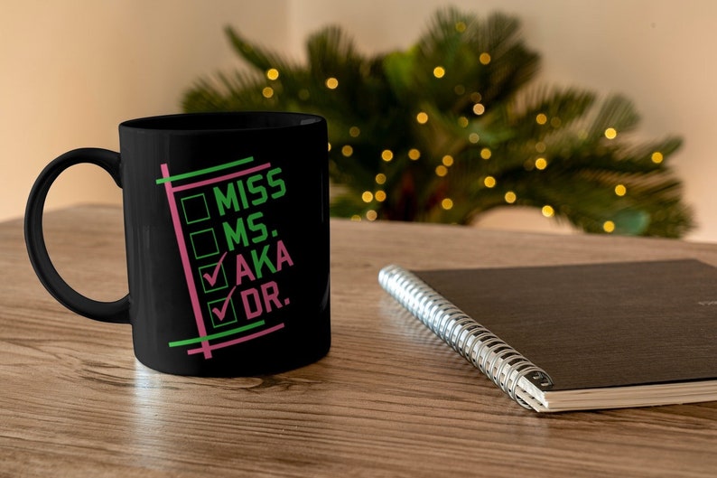 Puede incluir: Taza de cer&aacute;mica negra con asa blanca. La taza presenta las palabras "MISS, MS., AKA, DR." en verde y rosa, con marcas de verificaci&oacute;n junto a "AKA" y "DR.". Un cuaderno y un &aacute;rbol de Navidad est&aacute;n en el fondo.