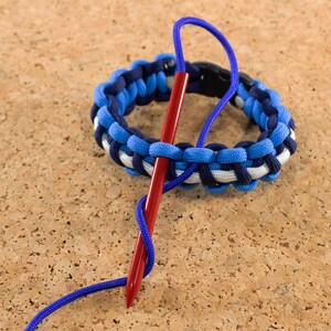 May include: A blue, white, and black paracord bracelet with a red needle threaded through it. The bracelet is being tied with a knot.