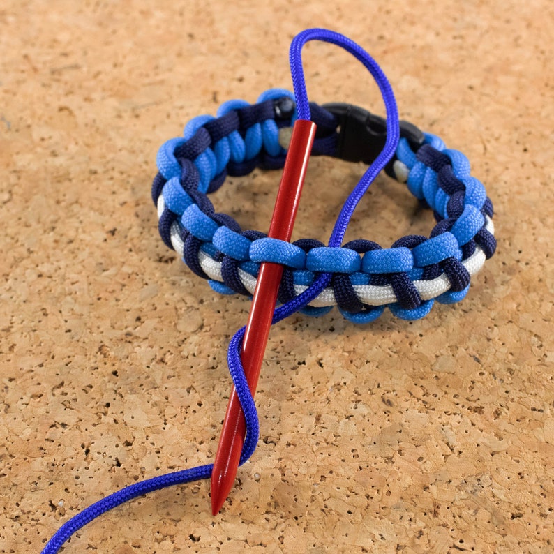 May include: A blue, white, and black paracord bracelet with a red needle threaded through it. The bracelet is being tied with a knot.