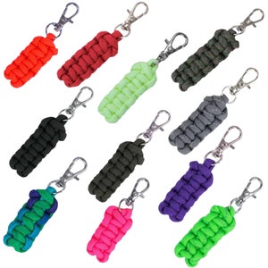 Könnte beinhalten: Zwölf bunte Paracord-Schlüsselanhänger mit silbernen Schlüsselringen. Die Schlüsselanhänger sind aus geflochtenem Paracord gefertigt und in verschiedenen Farben erhältlich, darunter Rot, Grün, Blau, Schwarz und Pink.