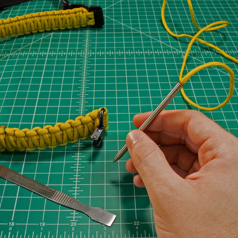 May include: A close-up of a hand holding a metal awl, a tool used for piercing holes in leather or other materials. The awl is resting on a green cutting mat with a yellow paracord bracelet and a small metal tool.