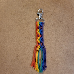 May include: A handmade macrame keychain featuring a rainbow color scheme. The keychain has a silver-colored clasp and a woven design in red, orange, yellow, green, blue, and purple. The bottom of the keychain has a fringe of the same rainbow colors.