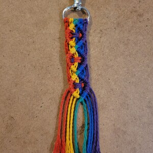 May include: A handmade macrame keychain with a rainbow color scheme. The keychain features a silver-colored clasp and a braided design in red, orange, yellow, green, blue, and purple. The bottom of the keychain has several strands of the same colors.