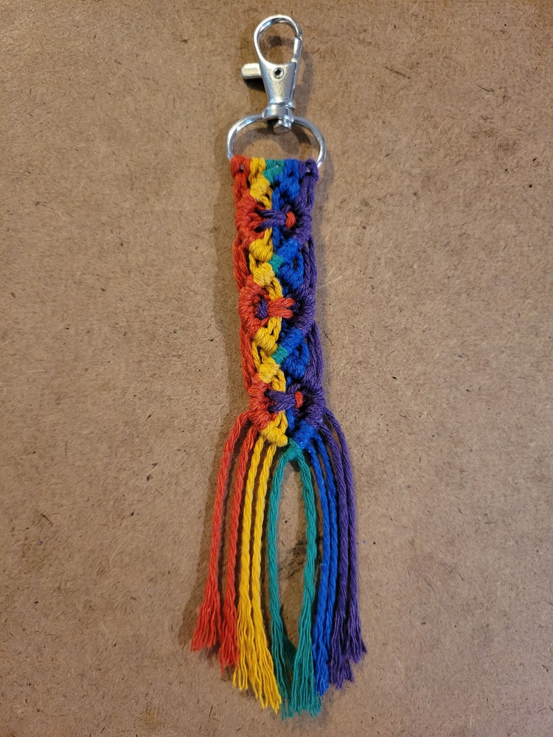 May include: A handmade macrame keychain with a rainbow color scheme. The keychain features a silver-colored clasp and a braided design in red, orange, yellow, green, blue, and purple. The bottom of the keychain has several strands of the same colors.