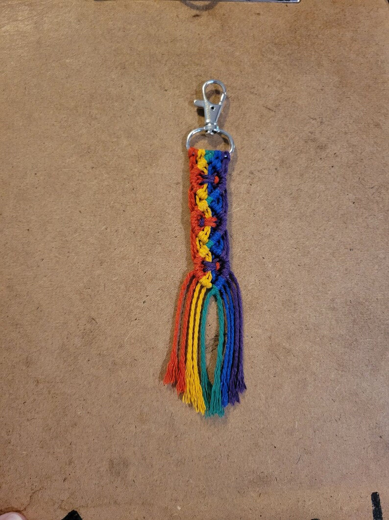 May include: A handmade macrame keychain featuring a rainbow color scheme. The keychain has a silver-colored clasp and a woven design in red, orange, yellow, green, blue, and purple. The bottom of the keychain has a fringe of the same rainbow colors.