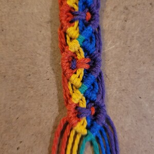 May include: A handmade macrame keychain featuring a rainbow color scheme. The keychain has a braided design with red, orange, yellow, green, blue, and purple threads. A silver metal ring is attached at the top.