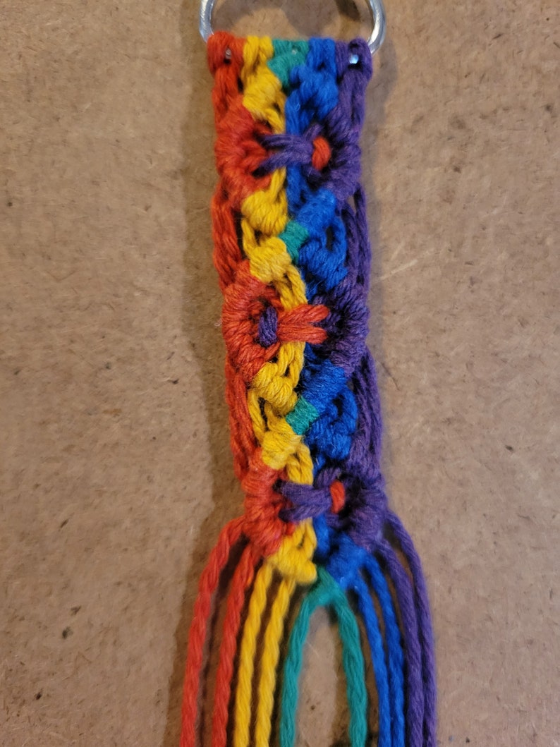 May include: A handmade macrame keychain featuring a rainbow color scheme. The keychain has a braided design with red, orange, yellow, green, blue, and purple threads. A silver metal ring is attached at the top.