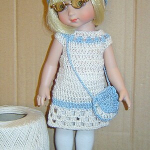 Könnte beinhalten: Eine Puppe, die ein weißes und blaues Häkelkleid mit einer passenden blauen Häkeltasche trägt. Die Puppe hat blonde Haare, blaue Augen und trägt eine Brille. Die Puppe trägt weiße Strumpfhosen und schwarze Schuhe.
