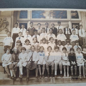 May include: Sepia-toned photograph of a class of students posing on steps in front of a building. The students, a mix of boys and girls, are dressed in various outfits, including shirts, ties, and dresses. The image is a vintage school portrait.