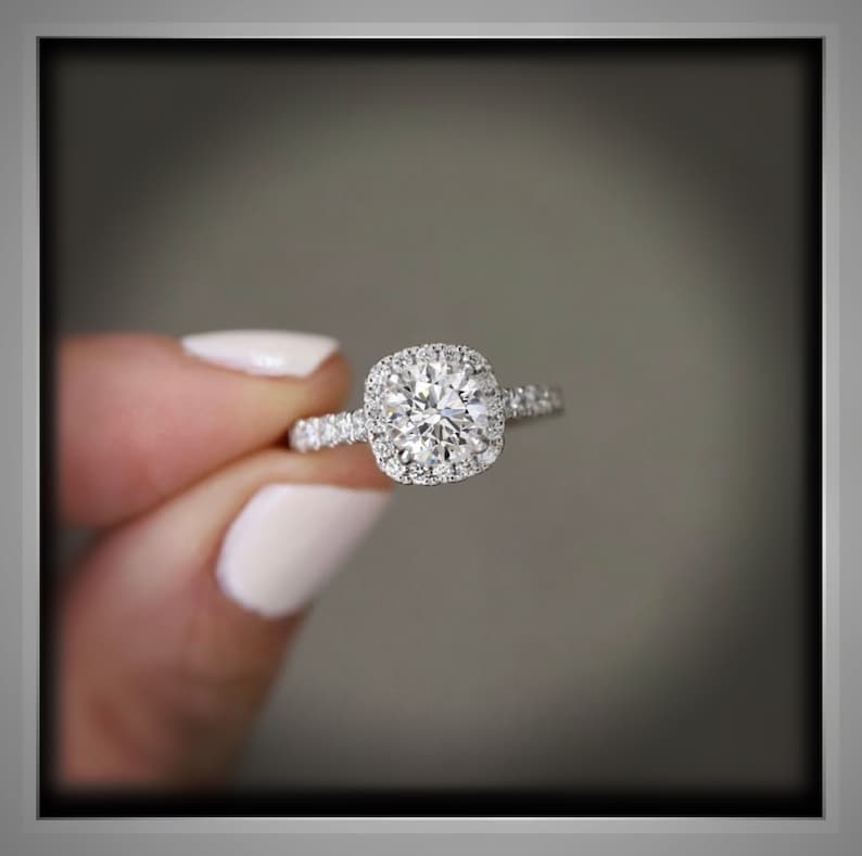 K&ouml;nnte beinhalten: Ein Diamant-Verlobungsring mit einem runden Mittelstein und einem Halo aus kleineren Diamanten. Das Band des Rings ist ebenfalls mit kleinen Diamanten besetzt. Der Ring wird vor einem verschwommenen Hintergrund gehalten.