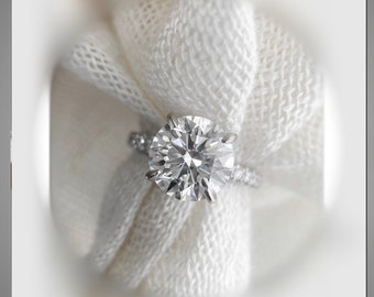 May include: A close-up of a sparkling diamond engagement ring set in a white gold band. The ring is resting on a white fabric background.