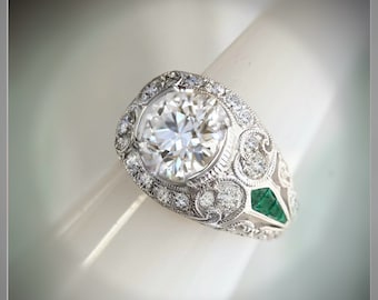 May include: A silver ring with a large round diamond center stone surrounded by smaller diamonds. The ring has a detailed design with emerald green stones on the side.