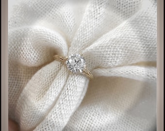 May include: A gold engagement ring with a large, round, brilliant-cut diamond set in a delicate, prong setting. The ring is on a white fabric background.