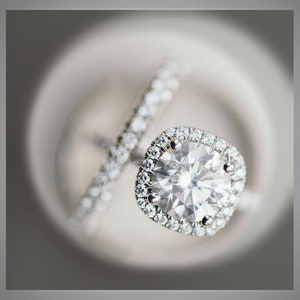 K&ouml;nnte beinhalten: Nahaufnahme eines Diamant-Verlobungsrings mit Halo-Fassung und passendem Band. Der Ring hat einen gro&szlig;en, rund geschliffenen Diamanten, der von kleineren Akzentdiamanten umgeben ist. Das Band ist ebenfalls mit kleinen Diamanten besetzt.
