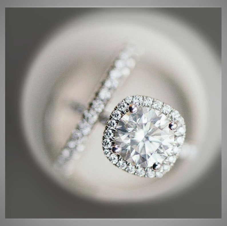 K&ouml;nnte beinhalten: Nahaufnahme eines Diamant-Verlobungsrings mit Halo-Fassung und passendem Band. Der Ring hat einen gro&szlig;en, rund geschliffenen Diamanten, der von kleineren Akzentdiamanten umgeben ist. Das Band ist ebenfalls mit kleinen Diamanten besetzt.
