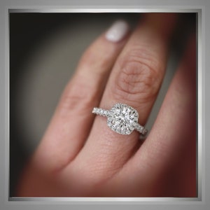 K&ouml;nnte beinhalten: Ein Diamant-Verlobungsring mit quadratischer Halo-Fassung. Der Ring hat einen gro&szlig;en, rund geschliffenen Mittelstein, der von kleineren Diamanten umgeben ist. Das Band ist ebenfalls mit Diamanten besetzt.