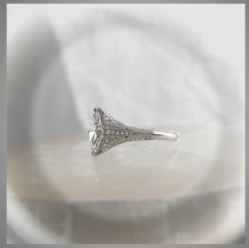 Pu&ograve; includere: Un anello di fidanzamento in argento con un grande diamante di forma ovale incastonato in un design delicato e intricato. L'anello &egrave; posto su una superficie bianca.