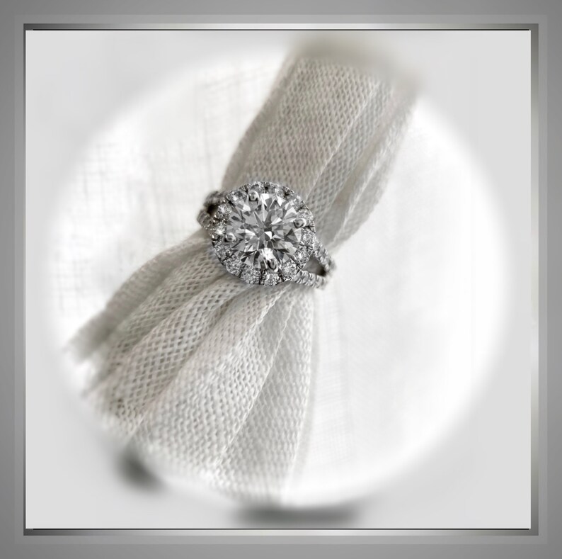 Op de afbeelding: Een zilveren verlovingsring met een grote, ronde, heldere diamant omgeven door een halo van kleinere diamanten. De ring ligt op een witte stoffen achtergrond.