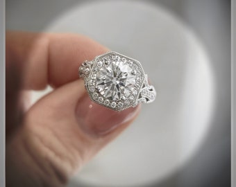 May include: A silver engagement ring with a large, round, clear diamond set in a hexagonal halo. The diamond is surrounded by smaller diamonds.