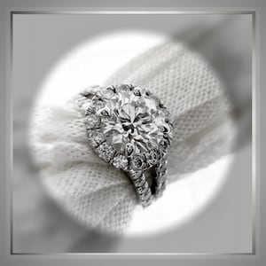 Op de afbeelding: Een close-up van een zilveren ring met een grote, ronde, glinsterende diamant omringd door kleinere diamanten. De ring ligt op een witte stoffen achtergrond.