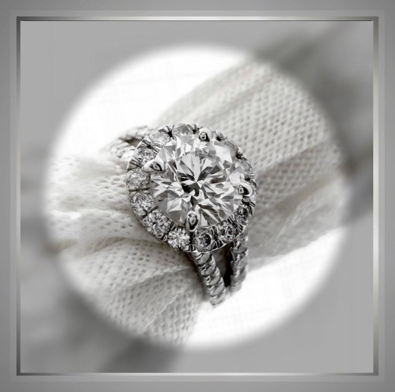 Op de afbeelding: Een close-up van een zilveren ring met een grote, ronde, glinsterende diamant omringd door kleinere diamanten. De ring ligt op een witte stoffen achtergrond.