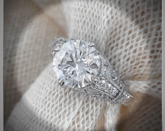 May include: A silver engagement ring with a large, round, brilliant-cut diamond surrounded by smaller diamonds. The ring is set on a white fabric background.