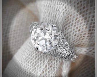 May include: A close-up of a diamond ring with a large, clear, oval-cut center stone. The ring is set in a detailed, ornate silver band with smaller accent stones. The ring is displayed on a white, textured fabric.