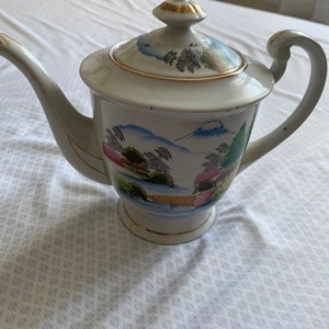 May include: Cream ceramic teapot with lid, spout, and handle. The teapot is decorated with a landscape scene featuring mountains, trees, and a bridge. Gold trim accents the edges of the lid, spout, and base. The teapot has a classic, vintage design.