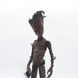 May include: A brown and green 3D printed figurine of a tree-like character with a human-like face. The character is holding a small, silver object in its right hand. The figurine is standing on a gray circular base.