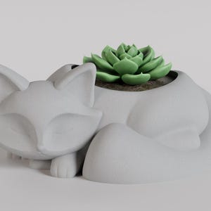 May include: A white ceramic planter shaped like a sleeping cat with a green succulent plant growing inside.