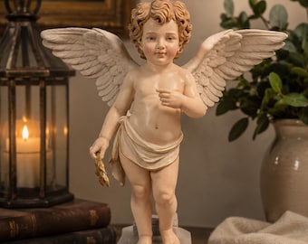 May include: A standing cherub figurine with curly light brown hair, large cream-coloured feathered wings, and a draped cloth around the waist. The figure holds a small golden branch in one hand and stands on a rectangular grey stone-like base.