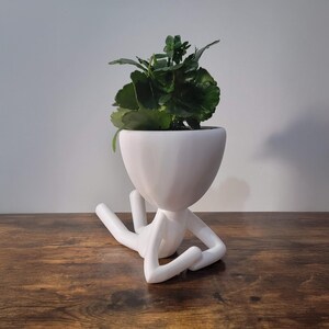 Puede incluir: Maceta de cerámica blanca con forma de persona tumbada con una planta verde en su interior.