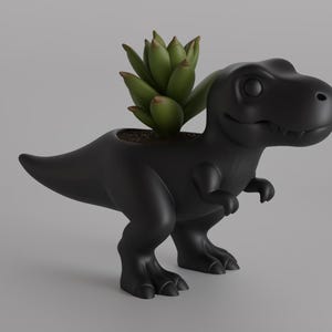 May include: A black Tyrannosaurus Rex planter with a green succulent plant. The dinosaur-shaped planter has a smiling face and is made of a matte black material. The plant is centered in the planter's head.