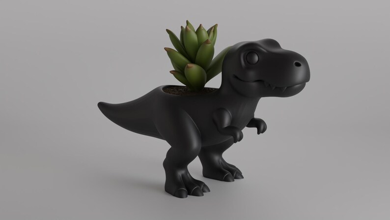 May include: A black Tyrannosaurus Rex planter with a green succulent plant. The dinosaur-shaped planter has a smiling face and is made of a matte black material. The plant is centered in the planter's head.