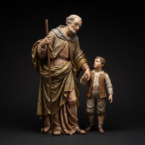 Puede incluir: Una escultura detallada representa una figura mayor con una venda en los ojos y un bastón, tomados de la mano de un niño. Las figuras visten túnicas y chalecos drapeados, con una paleta de colores apagados de marrones, dorados y cremas. La escultura está sobre un fondo oscuro.