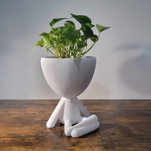 Könnte beinhalten: Weißer Keramik-Pflanztopf in Form einer sitzenden Person mit gekreuzten Beinen, die eine grüne Pflanze hält.