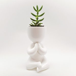 May include: White ceramic planter shaped like a person sitting in a yoga pose, holding a small green succulent plant in the head of the planter.