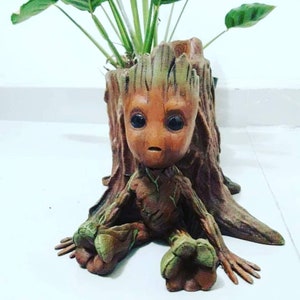 May include: A brown and green Groot planter with a plant growing out of the top. The planter is shaped like a tree stump and has a small Groot figure sitting on it.