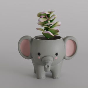 Puede incluir: Una maceta gris con forma de elefante con orejas internas rosas y una trompa. Contiene una planta suculenta verde y rosa. La maceta tiene ojos negros y mejillas rosas. El fondo es gris claro liso.