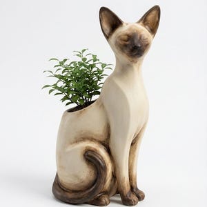 以下が含まれることがあります： 小さな緑の植物が入った、セラミック製のシャム猫のプランター。猫はクリーム色で、顔、耳、尾に茶色のアクセントがあります。猫は目を閉じて直立しており、植物は背中の空洞から生えています。