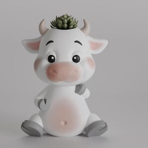 Puede incluir: Maceta de cerámica blanca en forma de vaca con una pequeña planta suculenta verde que crece en la parte superior de su cabeza. La maceta de vaca tiene una nariz y mejillas rosadas y está sentada sobre una superficie blanca.
