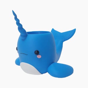 Puede incluir: Una maceta azul con forma de narval con un cuerno en espiral, ojos negros y mejillas rosadas. La maceta tiene una barriga blanca y una aleta caudal. El recipiente está diseñado para contener pequeñas plantas u otros objetos decorativos.