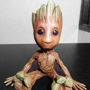 May include: A brown and green figurine of a character from the Guardians of the Galaxy movie. The character is sitting with its legs crossed and has a large head with a green leafy crown. The figurine is made of a textured material that looks like wood.